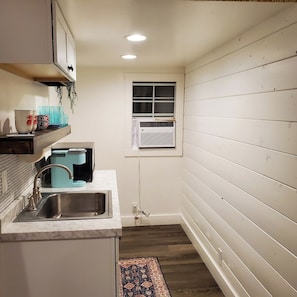 A compact kitchen with a sink, a window, and a built-in air conditioner. - Cozy Cottage on a Working Flower Farm — Private Deck, Fire Pit, Near Sacramento (Loomis)