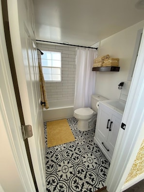 A bathroom with a white bathtub, a toilet, and a sink. There is a window above the toilet and a towel hanging on the door. - Cozy Cottage on a Working Flower Farm — Private Deck, Fire Pit, Near Sacramento (Loomis)