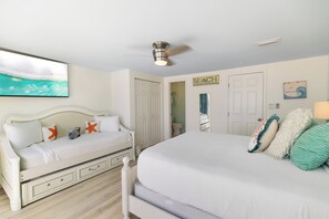 Ein Schlafzimmer mit einem Bett, einem Nachttisch, einer Kommode, einem Deckenventilator und einem Bild mit Strandmotiv.