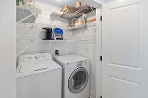 A laundry room with a front-loading washer and dryer, shelves above holding various items, and a door to the right. - Palapa House • Gulf Front • Palapa + Hammock • Sleeps 11 (Panama City Beach)