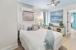 A bedroom with a bed, an air conditioning unit, a ceiling fan, a painting of a beach, a nightstand, a lamp, and a door leading to another room. - Palapa House • Gulf Front • Palapa + Hammock • Sleeps 11 (Panama City Beach)