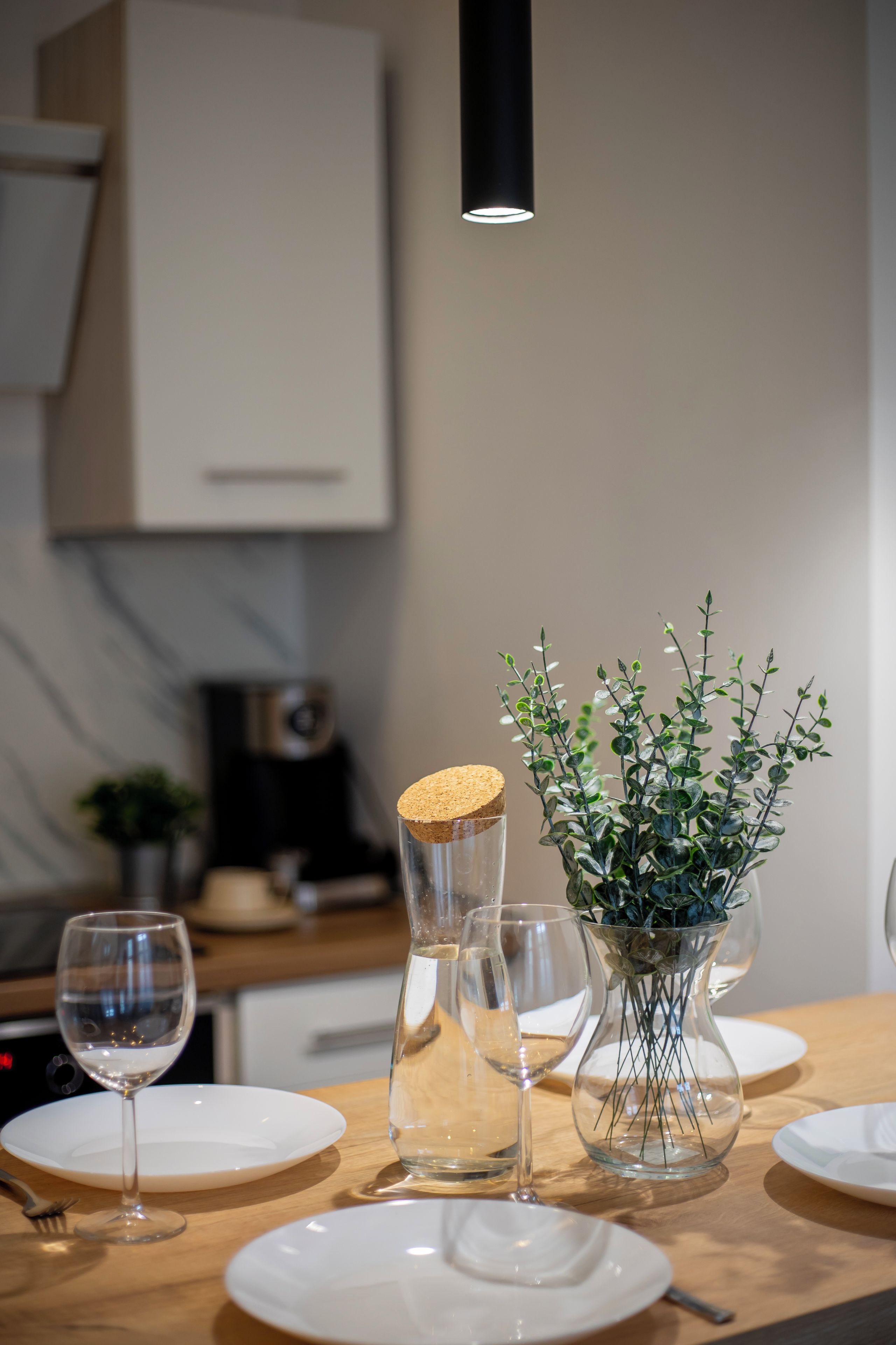 Una mesa de comedor con platos blancos, un florero de vidrio con follaje verde y una jarra de vidrio con tapón de corcho.