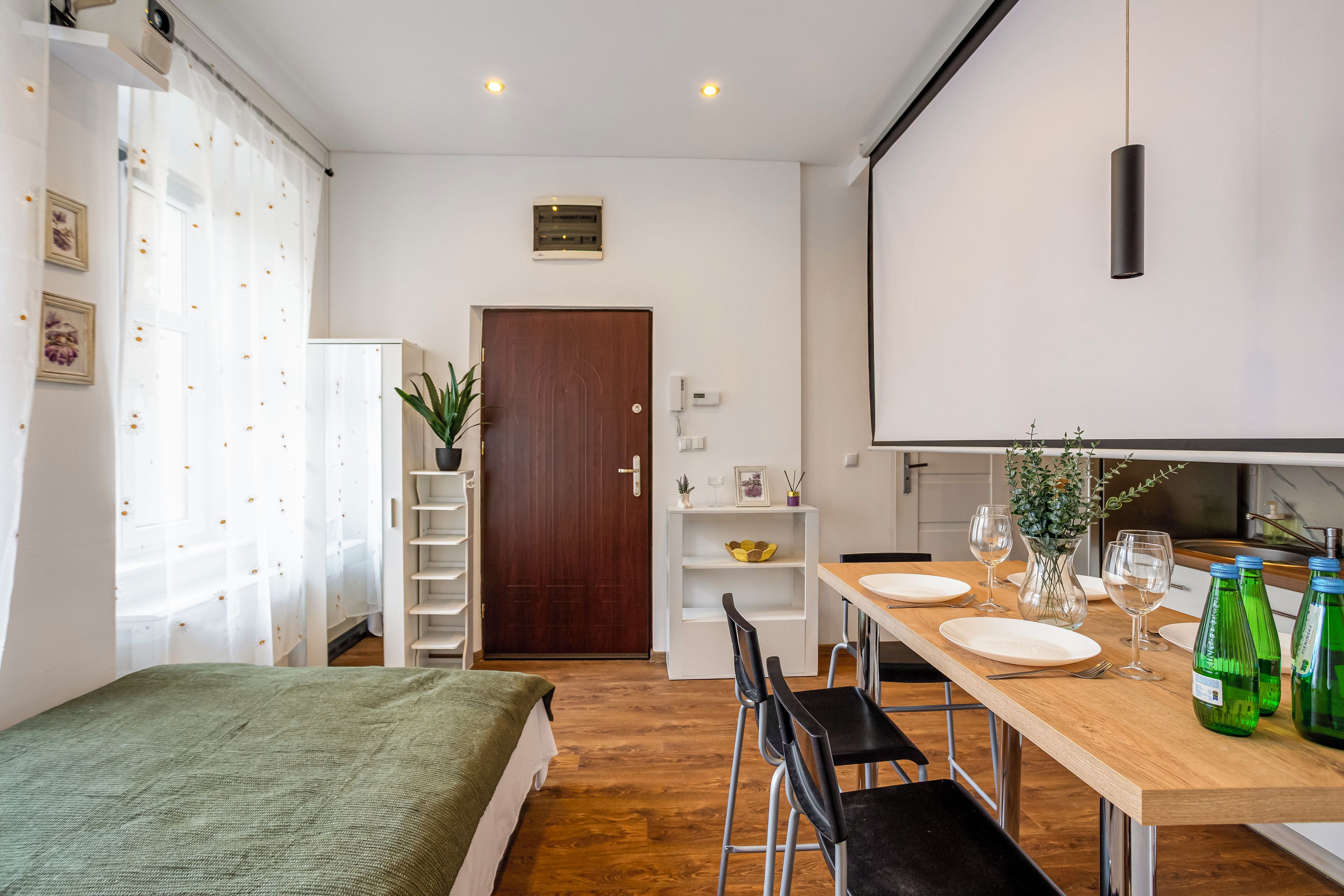 Habitación de hotel pequeña con cama, mesa de comedor para dos personas, una cocina compacta y una puerta que da a otra habitación.