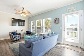 A living room with a blue sofa, patterned armchairs, a ceiling fan, a TV, and a door labeled 'BEACH HOUSE'. - A Kure for the Sole 5 bedroom 3 bathroom condo combo. With work from home space (Kure Beach)
