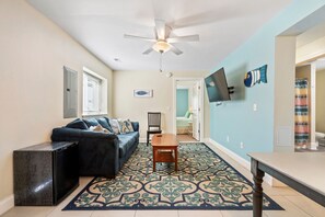 Ein Wohnzimmer mit einem blauen Sofa, einem Holztisch, einem Deckenventilator und einem an der Wand befestigten Fernseher.