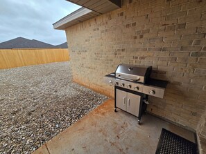 Ein Outdoor-Barbecue-Grill an einer Ziegelwand, mit Kiesfläche und einem Holenzaun im Hintergrund.