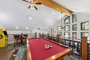 Une table de billard avec des billes, un ventilateur de plafond et une vue sur l’extérieur grâce à de grandes fenêtres.