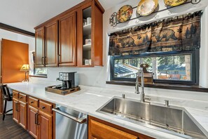 A kitchen with wooden cabinets, a stainless steel sink, and a window with a view of a house. - Lake & Après Chalet BBQ, Games, Pool Table (South Lake Tahoe)