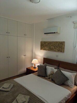 Een slaapkamer met een bed, kussens, een bedlampje, een teddybeer, een wandairconditioner en een kledingkast.