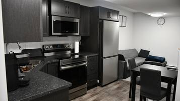 A modern kitchen with dark cabinetry, stainless steel appliances, a built-in oven, and a refrigerator. There is a dining area with a table and chairs.