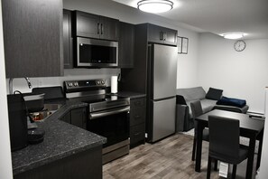A modern kitchen with dark cabinetry, stainless steel appliances, a built-in oven, and a refrigerator. There is a dining area with a table and chairs.