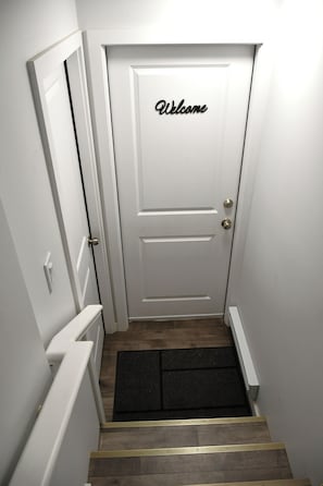 Un pasillo estrecho con una puerta blanca que tiene un letrero de 'Welcome', un felpudo y un pasamanos a la derecha.