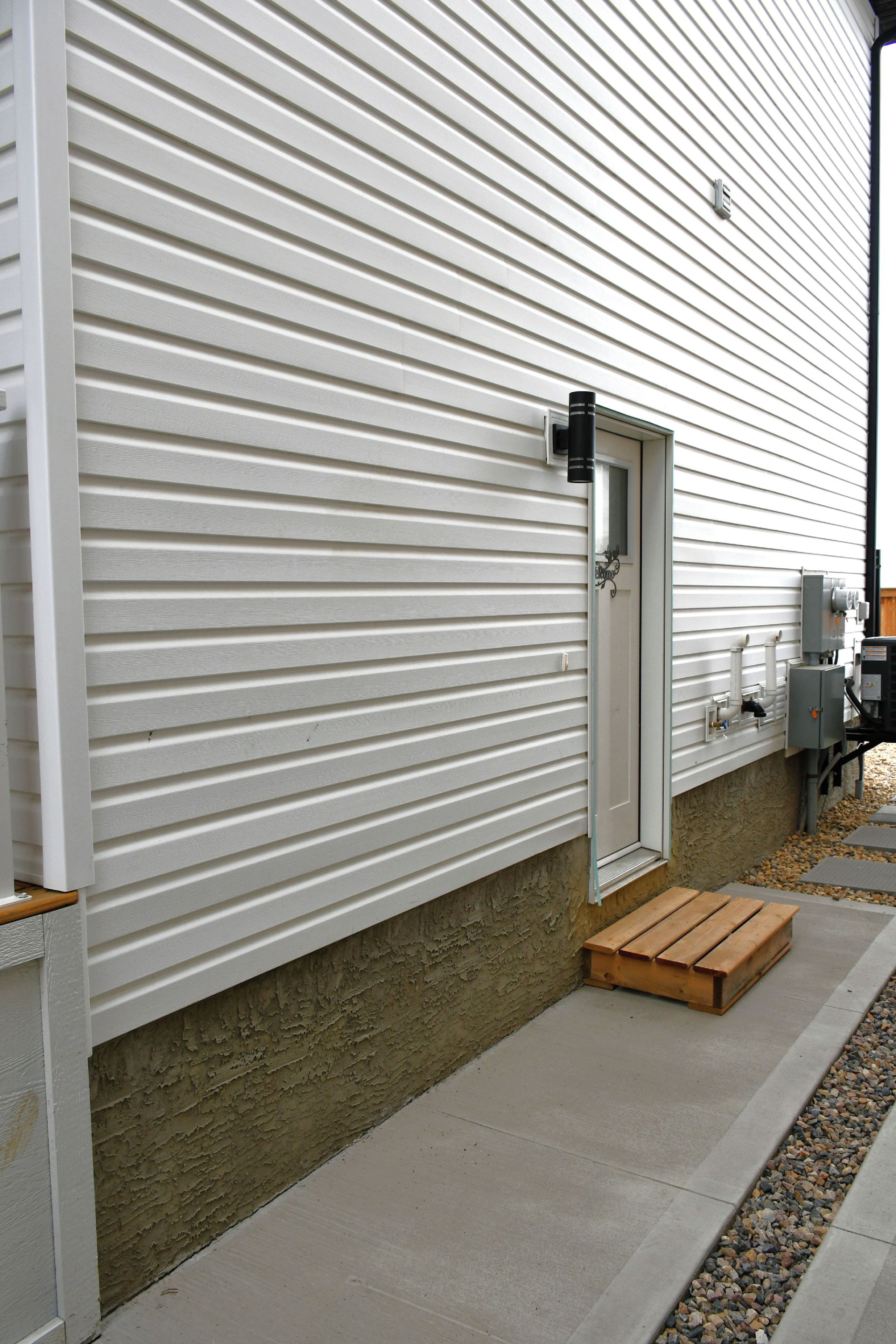 A modern exterior with a white siding wall, a metal door, and a concrete pathway leading to a wooden bench.