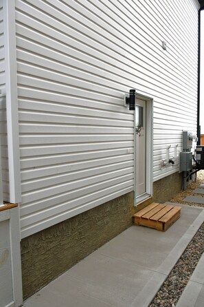 A modern exterior with a white siding wall, a metal door, and a concrete pathway leading to a wooden bench.