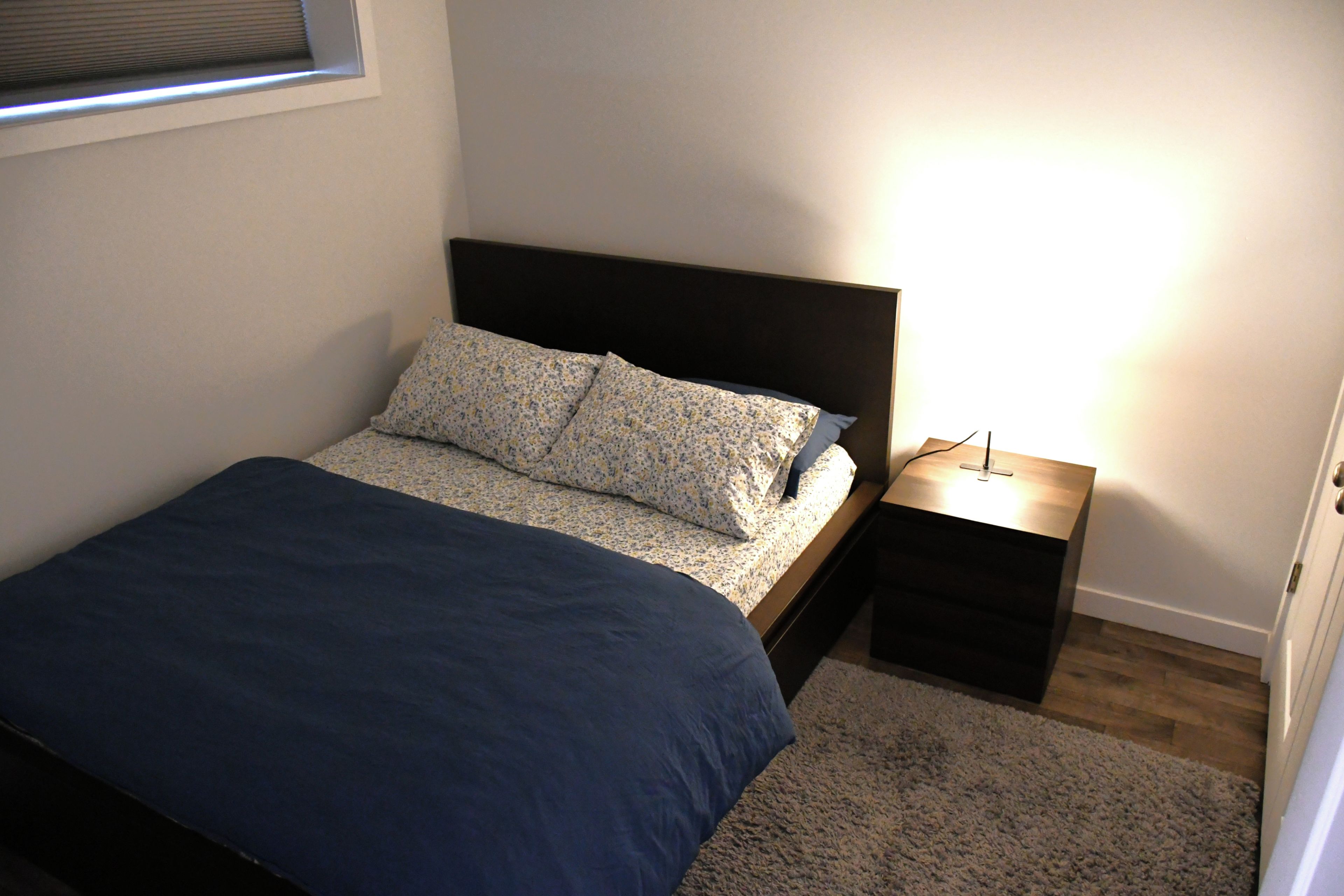 Ein Schlafzimmer mit einem Bett, einem Nachttisch, einer Lampe und einem Fenster.