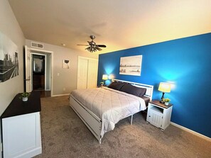 Ein Schlafzimmer mit einem Bett, Nachttischen, einem Deckenventilator, einer Tür und einer Wand mit einem Gemälde.