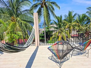 Eine Hängematte und ein farbenfroher Stuhl auf einer gefliesten Terrasse mit Palmen und Blick auf das Meer.