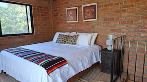 A bedroom with a brick wall, a bed with white bedding, a colorful blanket, and two framed pictures. - Lovely 1-bedroom house, steps from the beach and restaurants, with beach views. (Yelapa)