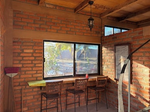 A room with a brick wall, a large window, and a wooden ceiling. There are two wicker chairs and a small table. - Lovely 1-bedroom house, steps from the beach and restaurants, with beach views. (Yelapa)