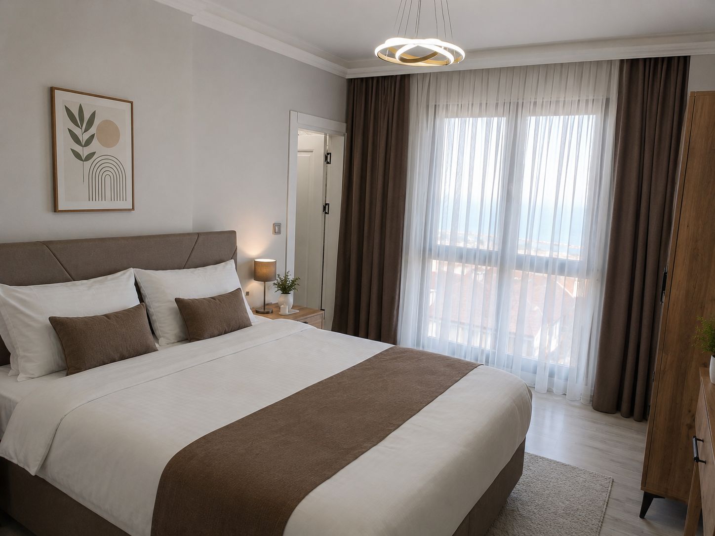 Une chambre d’hôtel avec un grand lit, une table de chevet avec une lampe, une vue sur l’extérieur à travers des rideaux légers et une œuvre d’art encadrée au mur.
