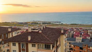 A residential area with multi-story buildings, a road with parked cars, and a view of the sea at sunset.