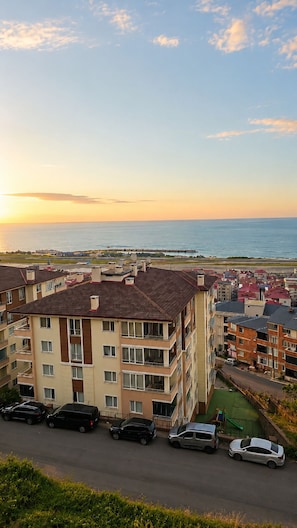 A residential area with multi-story buildings, a road with parked cars, and a view of the sea at sunset. - Airport Deluxe Suites (Trabzon)