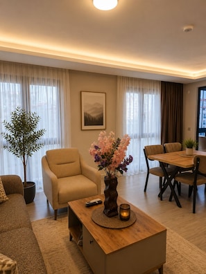 A modern living room with a sofa, armchair, dining table, and chairs. There is a potted plant and a vase with flowers on the coffee table. - Airport Deluxe Suites (Trabzon)