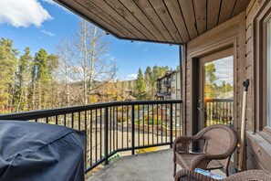 A balcony with a view of a forest and a building, a covered grill, and wicker furniture. - Walk to Lifts & Main St | Hot Tub (Breckenridge)