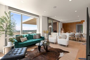 A modern living room with a green sofa, white armchairs, and a dining area with a wooden table and chairs. - Mosman Penthouse | Harbour Views + Parking (Mosman)