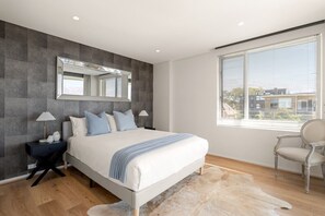Quarto moderno com cama grande, mesa de cabeceira, cadeira, janela com persianas e espelho na parede.