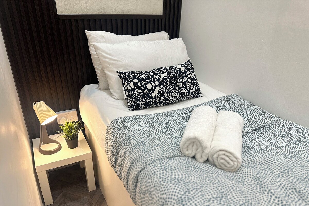A neatly made bed with white and patterned pillows, a blue patterned bedspread, and rolled towels on the bed.