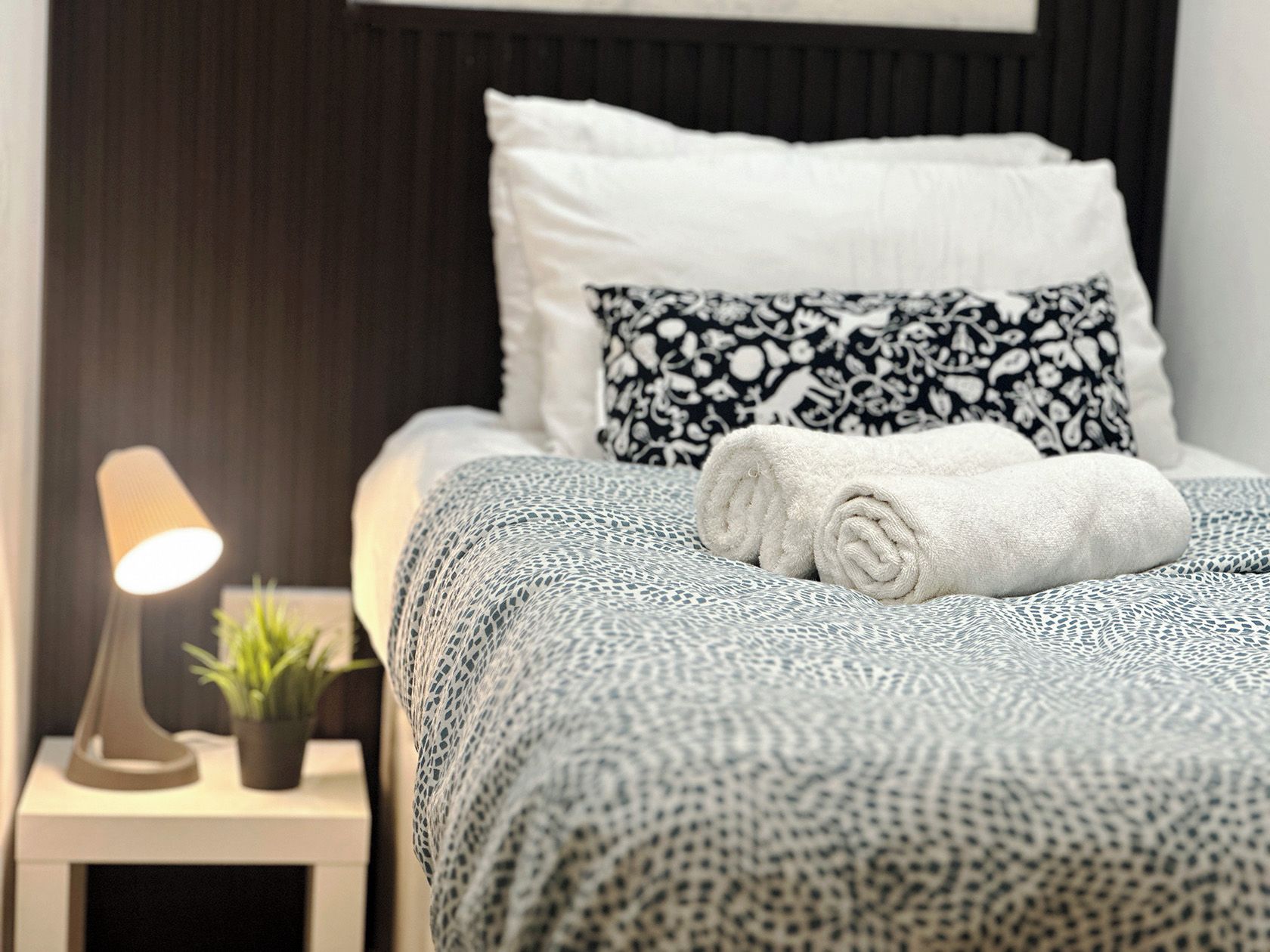 Een net opgemaakt bed met een bedsprei met een patroon, een opgerolde witte handdoek en een zwart-wit gebloemd kussen. Een bedlampje en een kleine potplant staan er ook.