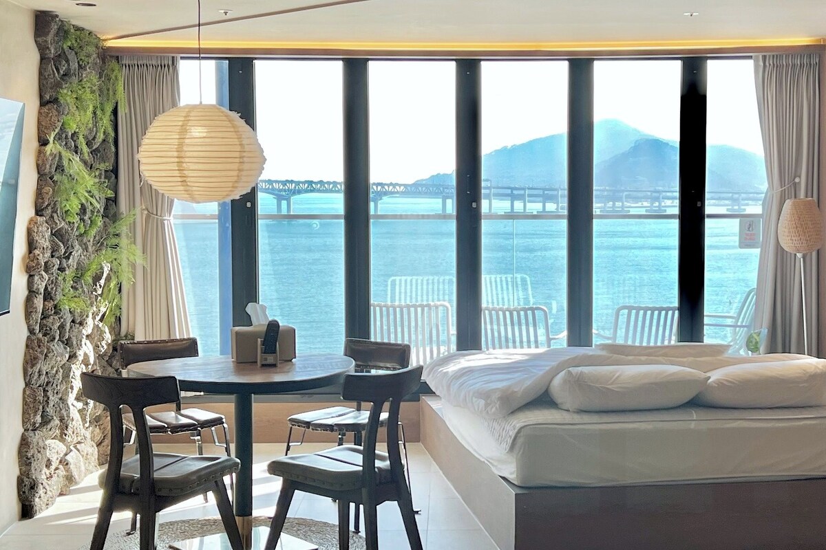 Un dormitorio con cama, una mesa de comedor con sillas, vista al mar y una pared de piedra.