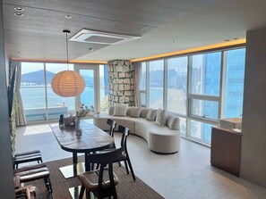A modern living room with a large window, a stone accent wall, a sofa, and a dining area with chairs and a table. - Lumiere (Busan)