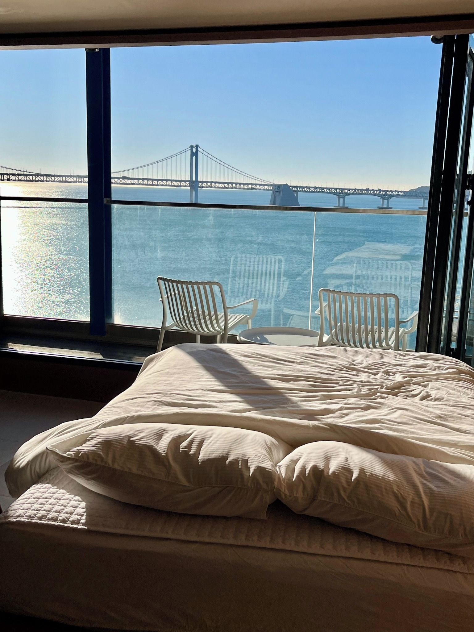 A bedroom with a large window overlooking a bridge and water. There is a bed with white bedding and two white chairs.