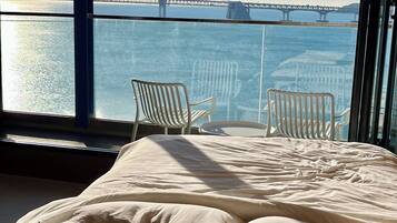 A bedroom with a large window overlooking a bridge and water. There is a bed with white bedding and two white chairs.