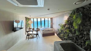 A modern bedroom with a bed, a dining table, and chairs, a TV mounted on the wall, and a view of the sea. - Lumiere (Busan)