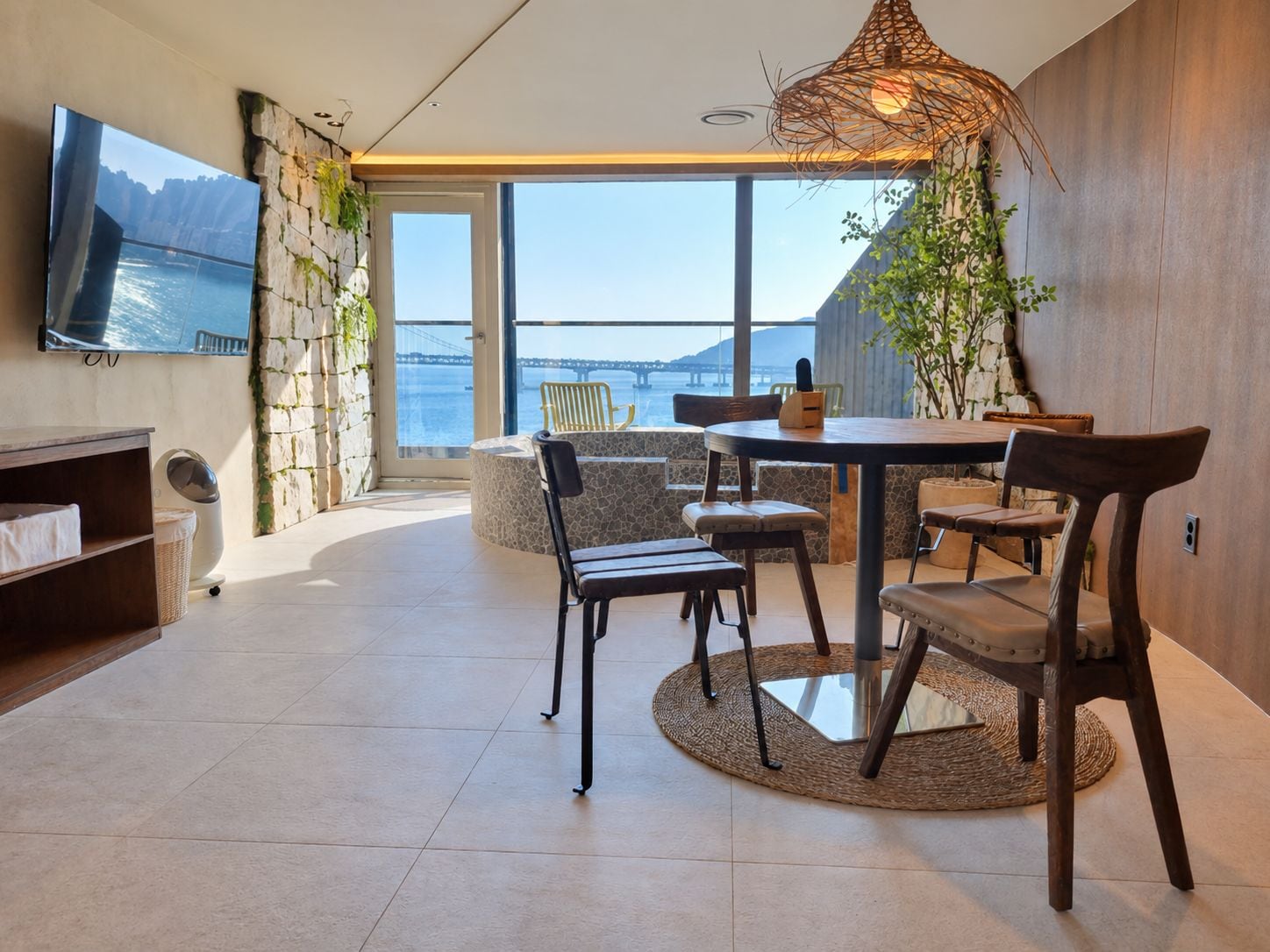 A modern interior with a dining area, a TV mounted on the wall, and a view of the sea through large windows.