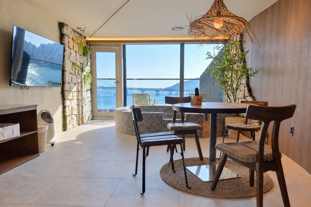 Un interior moderno con zona de comedor, televisor montado en la pared y vista al mar a través de amplias ventanas.