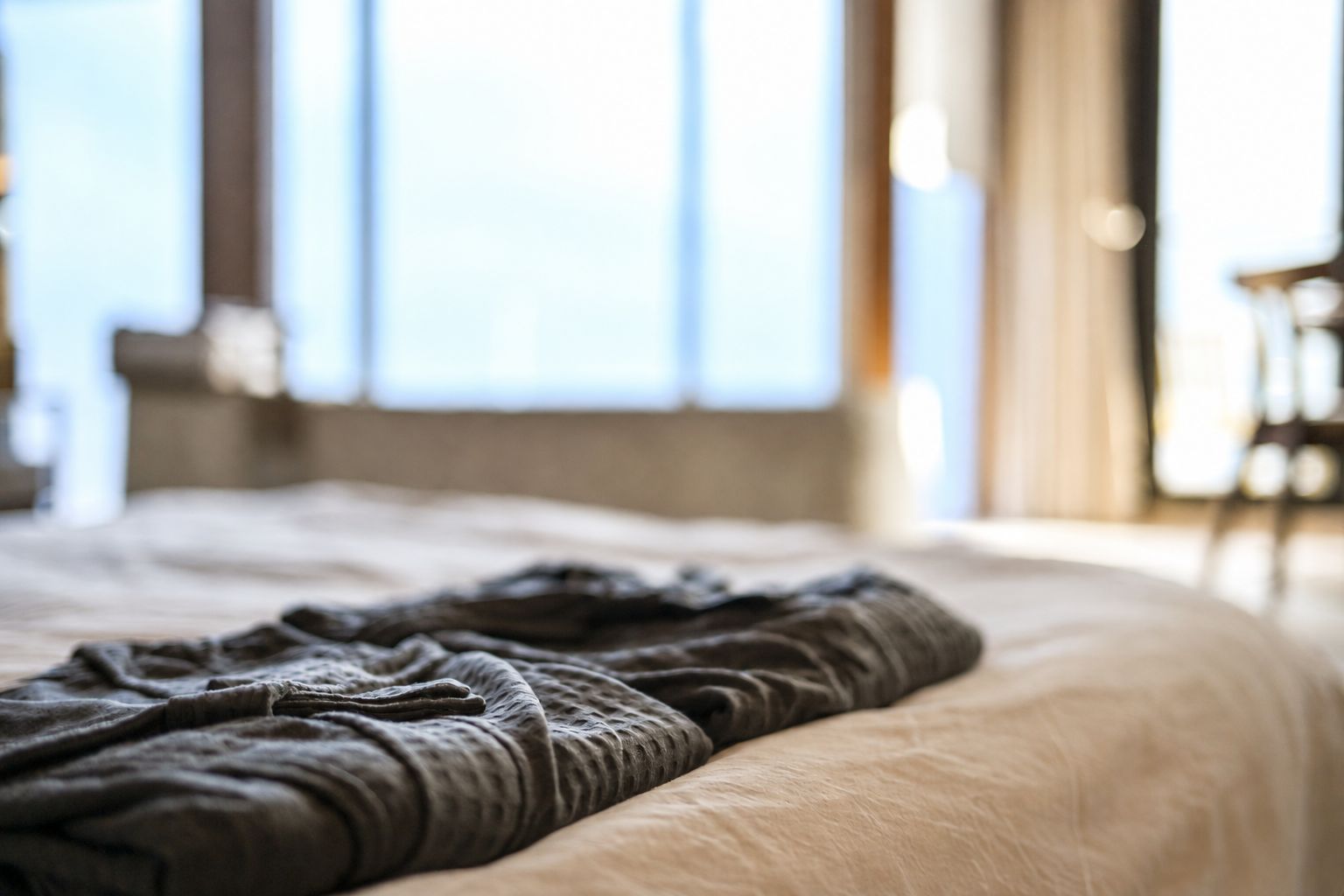 Een bed met een deken, een raam met gordijnen en een stoel in een kamer.