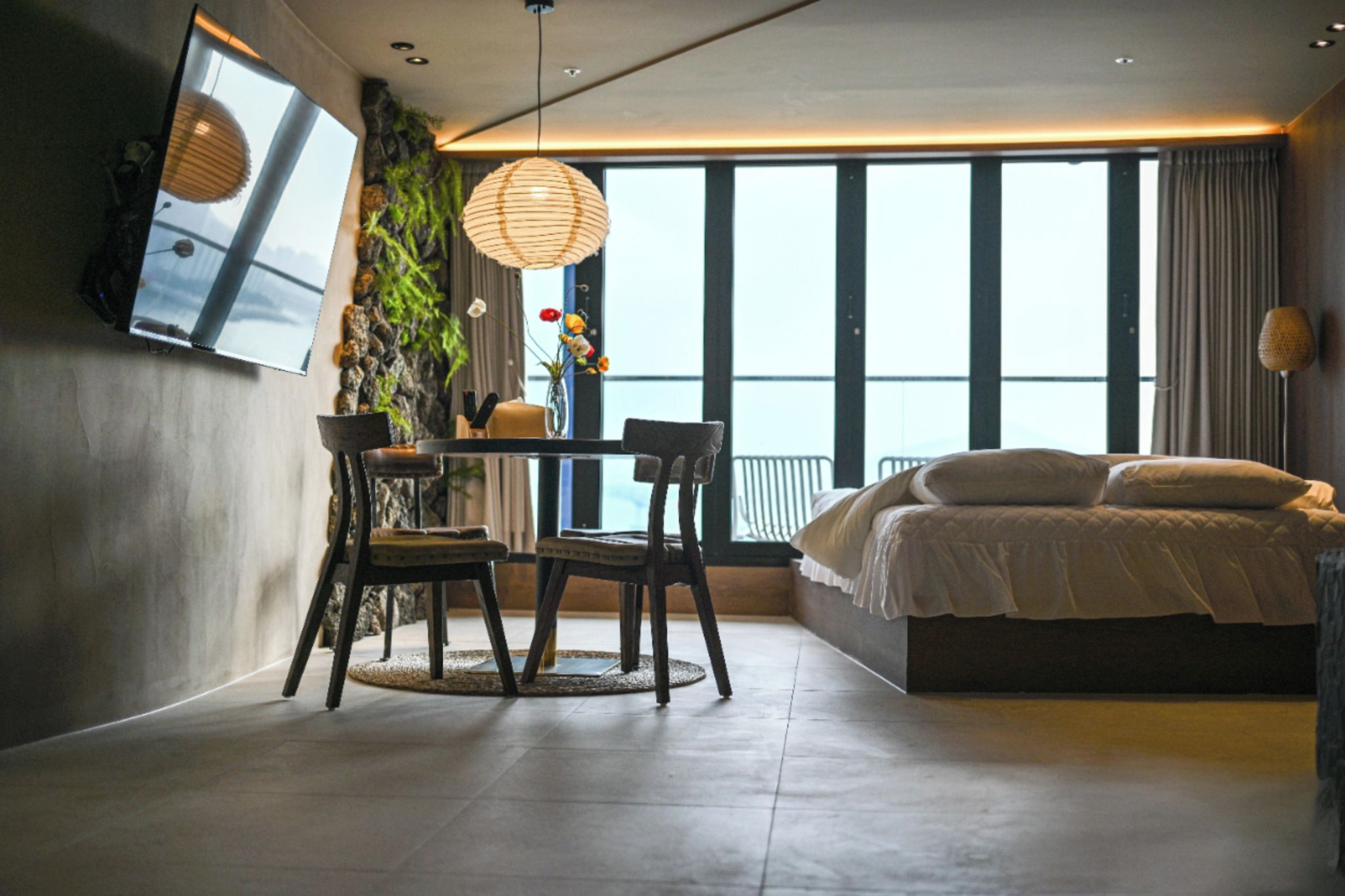 A modern bedroom with a large bed, a dining table with chairs, a hanging light fixture, and a view of the outdoors.