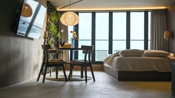 A modern bedroom with a large bed, a dining table with chairs, a hanging light fixture, and a view of the outdoors.