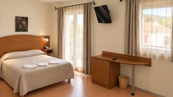 A hotel room with a bed, bedside table, lamp, TV mounted on the wall, and a desk with a chair.