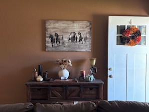 A room with a brown wall, a white door with a wreath, a wooden dresser with various items, and a painting of horses. - 3 bedroom home with hot tub and garage in Hays, KS (Hays)