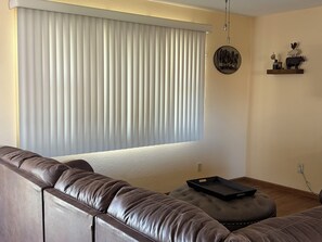 A living room with a brown leather sofa, a window with vertical blinds, a wall-mounted shelf with decorative items, and a power outlet. - 3 bedroom home with hot tub and garage in Hays, KS (Hays)