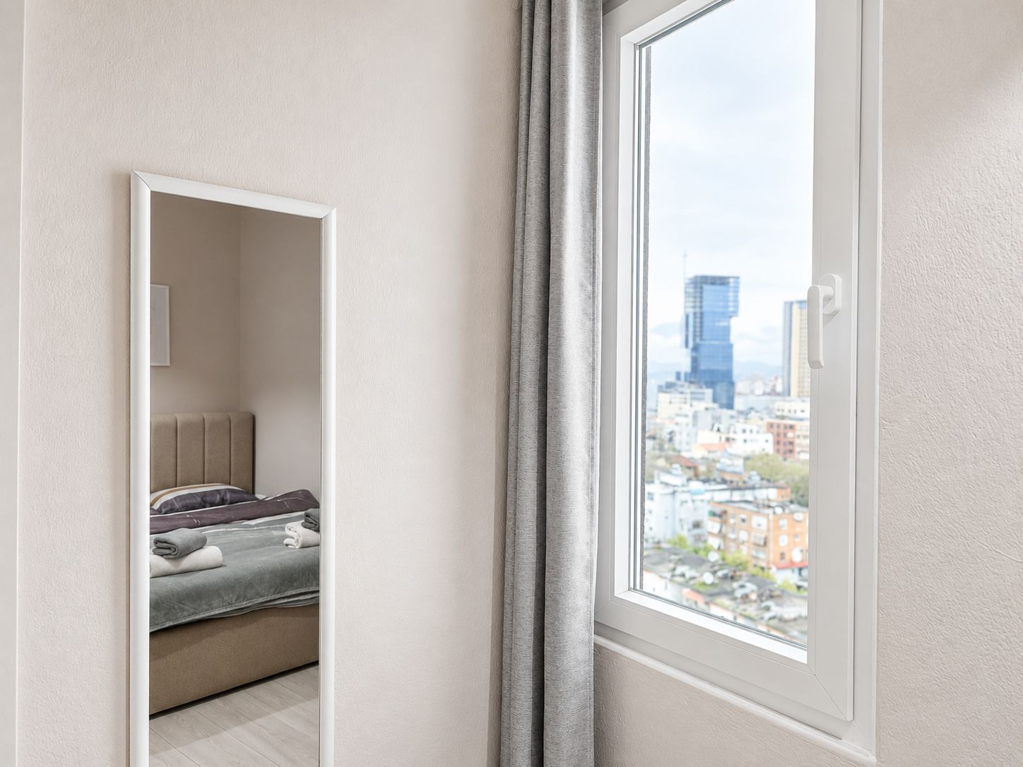 A bedroom with a bed, a bedside table, a window with a view of a cityscape, and a door leading to another room.