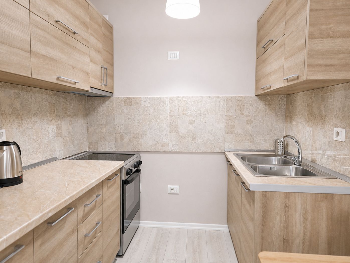 A compact kitchen with wooden cabinets, a built-in oven, a stainless steel sink, and a kettle on the countertop.
