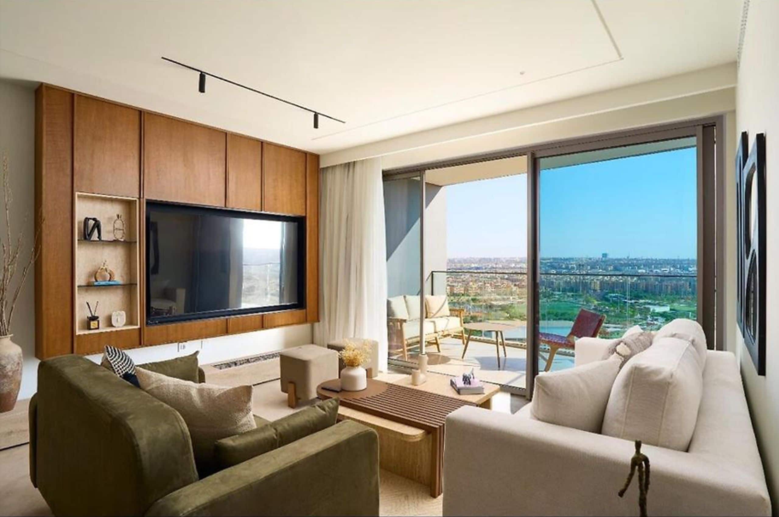 A modern living room with a large flat-screen TV, comfortable seating, and a balcony with a city view.