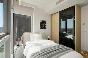Ein modernes Schlafzimmer mit einem Bett, einem Nachttisch, einer Lampe und Blick auf die Stadt durch ein großes Fenster.
