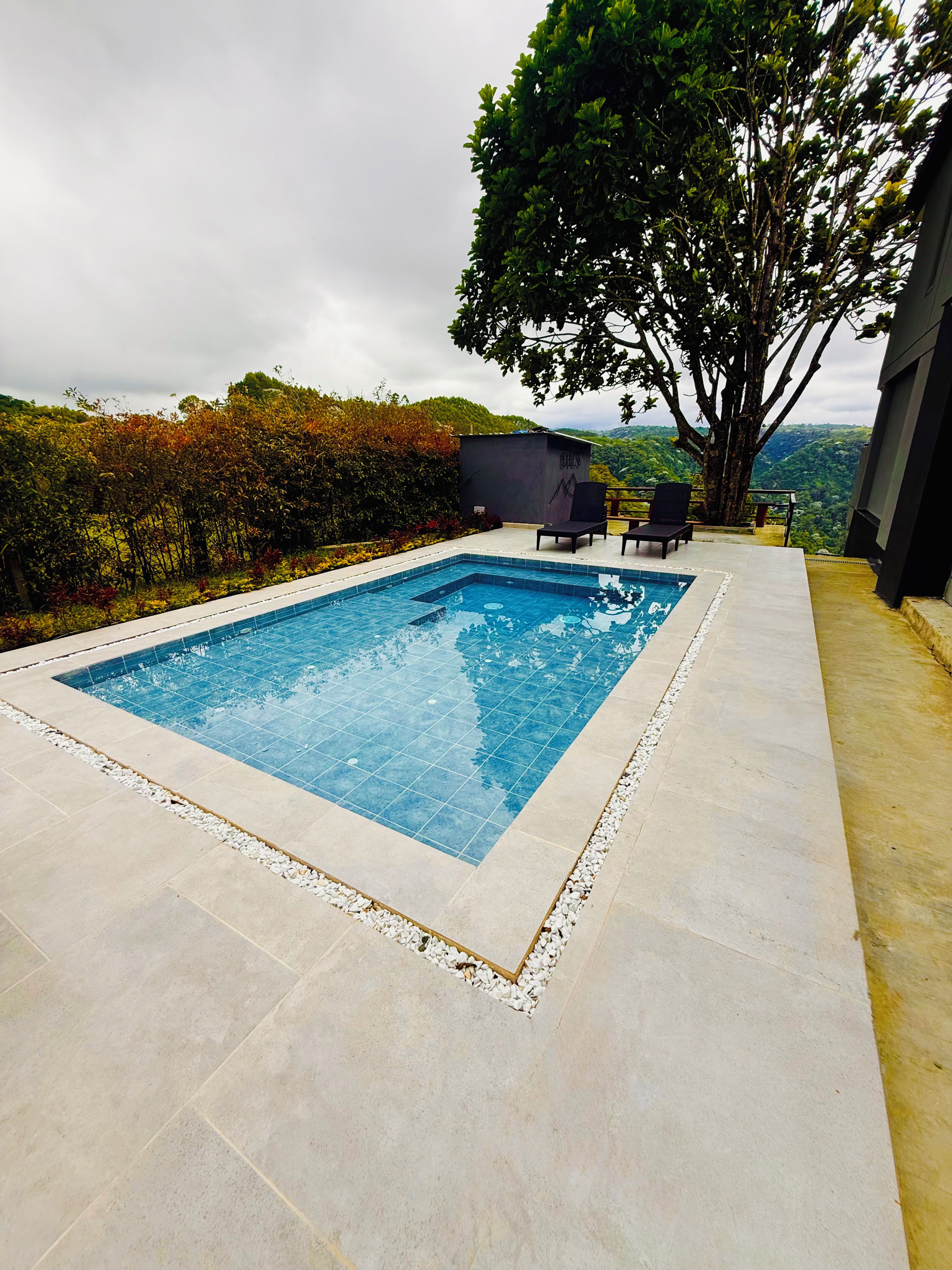 A rectangular swimming pool with a tiled edge, surrounded by a light-colored patio. There are two lounge chairs and a small table on the patio.